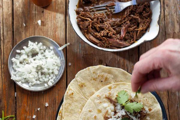 A person assembling a slow cooker barbacoa taco with a pot of shredded barbacoa meat, a small dish of onions, a bottle of hot sauce, and some cilantro around the taco plate.