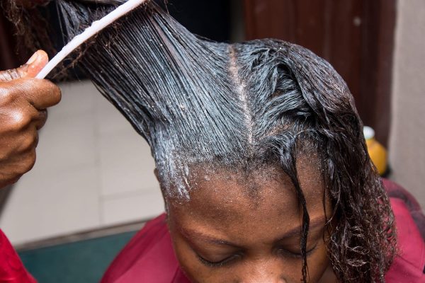 A photo of a hairdresser applying hair relaxer to her female client’s hair.