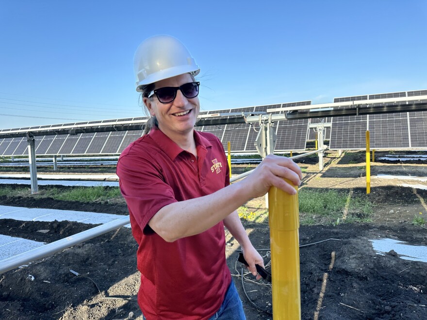 رجل يرتدي قبعة صلبة وقميصًا أحمر يقف أمام الألواح الشمسية.  مات أونيل، أستاذ علم الحشرات في جامعة ولاية أيوا، يستعرض مزرعة الطاقة الشمسية التجريبية التابعة للمدرسة جنوب أميس.