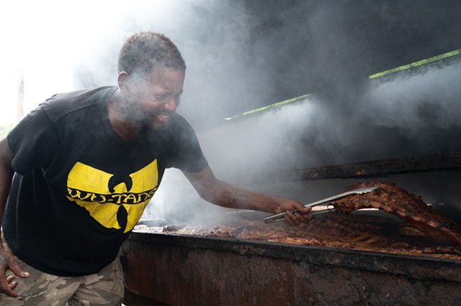 رجل ذو شعر قصير يرتدي قميص Wu-Tang يرفع رفًا متفحمًا من الضلوع من شواية مدخن