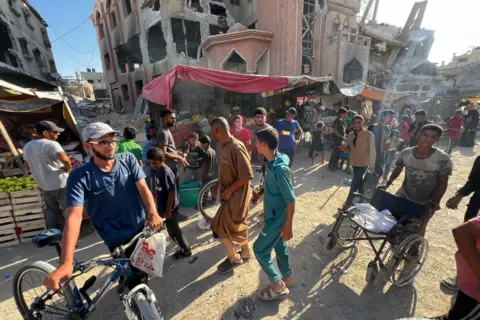 أشخاص أمام أكشاك في السوق في خان يونس، وخلفهم مباني مدمرة