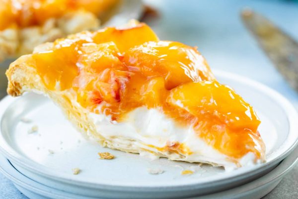 a photo of a single slice of creamy peach pie sitting on a small white dessert plate with a homemade crust, a creamy base layers and topped with a layer of juicy peaches.
