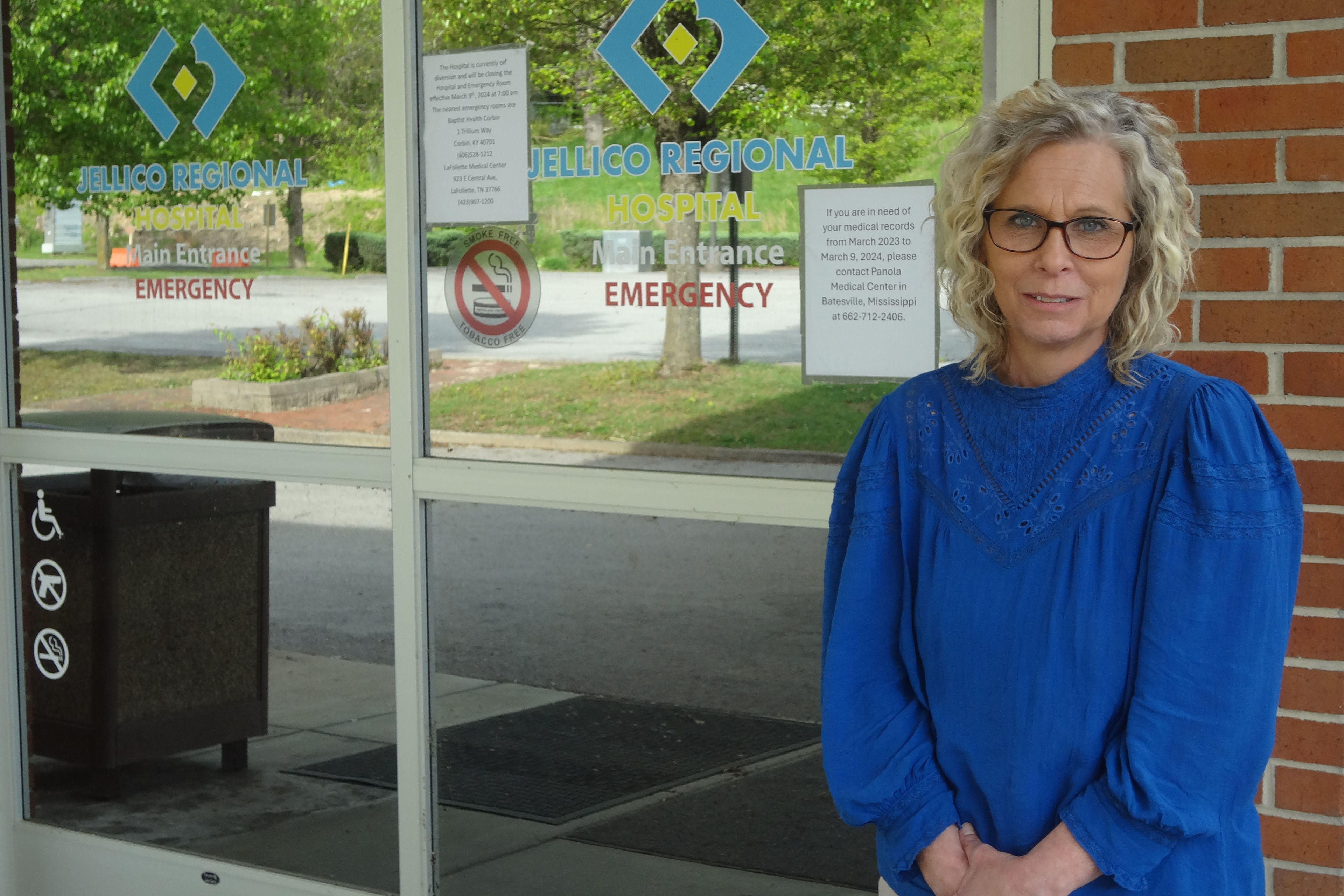 تقف ساندي تيري، التي ترتدي بلوزة زرقاء زاهية، بجانب الأبواب الزجاجية التي تمثل مدخل مركز جيليكو الطبي السابق.