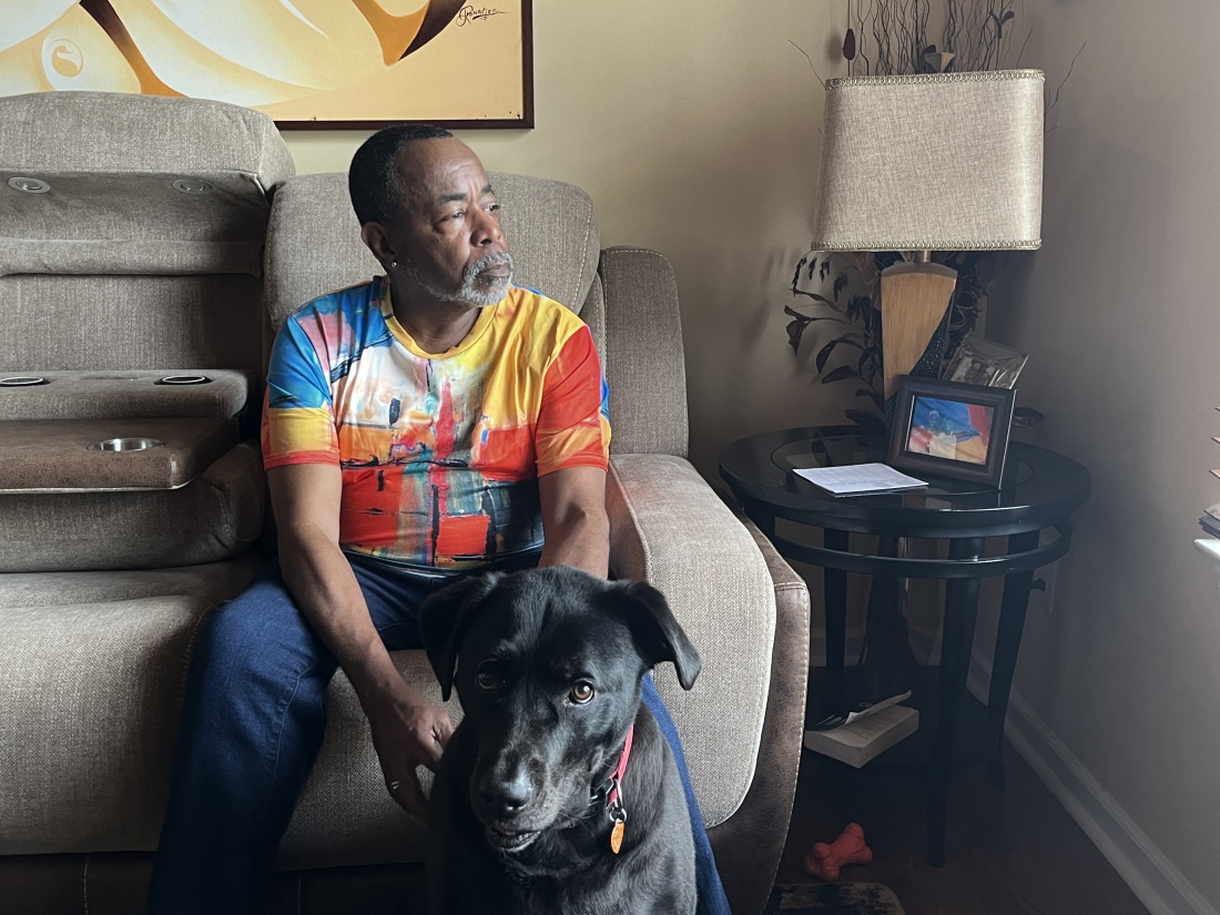 رجل يرتدي قميصًا ملونًا ولحية رمادية، يُدعى مالكولم ريد، يجلس على أريكة غرفة المعيشة ذات اللون البيج.  يدا ريد على كلبه، سامبسون، وهو مزيج من اللابرادور الأسود يجلس أمام الأريكة.  ينظر الكلب إلى الكاميرا بينما ينظر ريد نحو النافذة. 